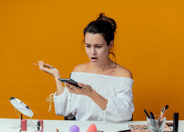 Screenshot 2025-12-16 at 09-46-02 Shocked beautiful girl sits at table with makeup tools looking at phone holding makeup brush isolated on orange wall Free Photo